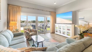 A spacious living room with a large television, comfortable seating, a glass door leading to an outdoor area, and a cabinet with various items. - Coastal Living Perfected: Banana Bay's Intracoastal Retreat with Sandy Shores (Pensacola)