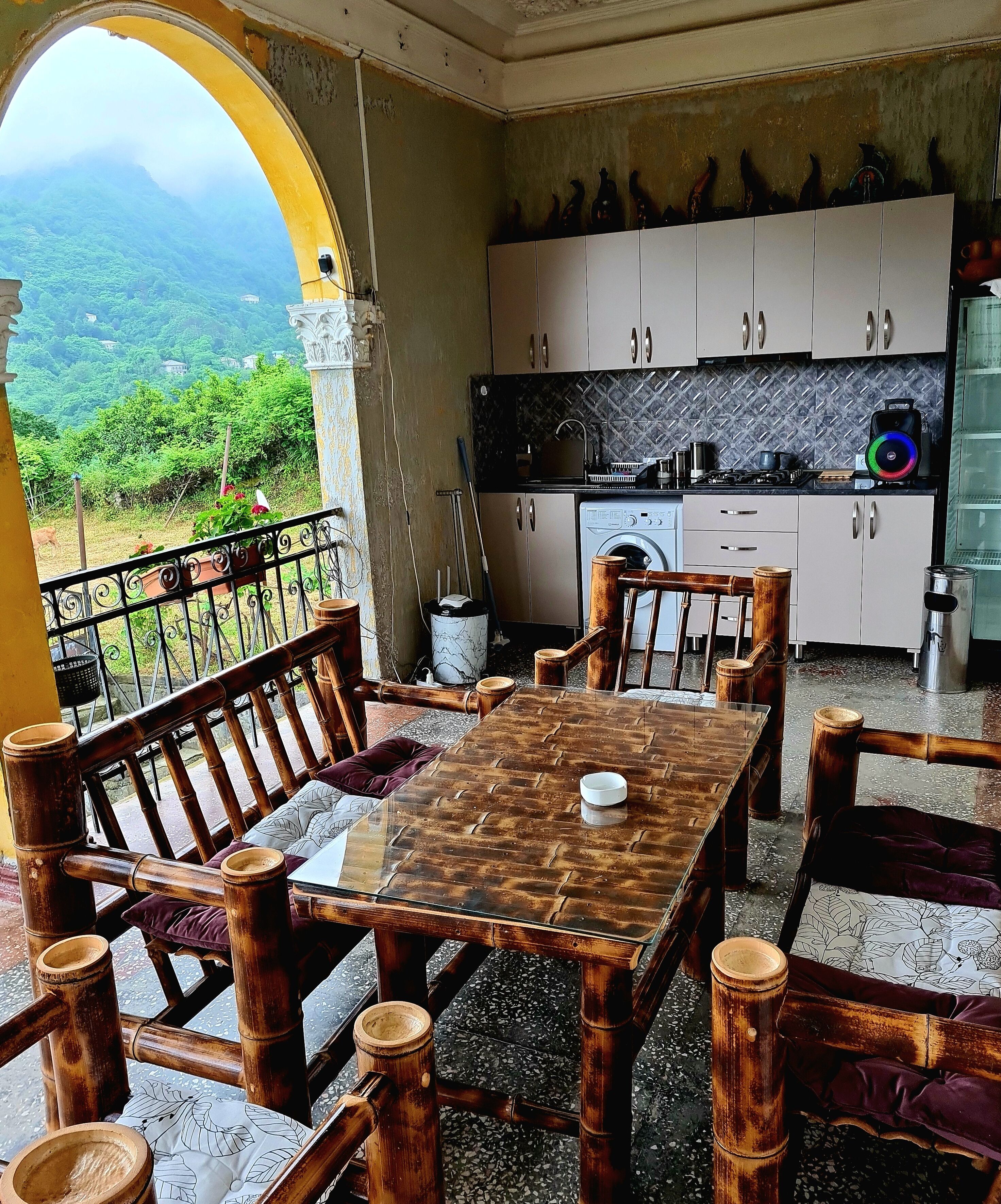 Un espace repas avec une table et des chaises, une cuisine équipée de placards et d’appareils électroménagers, et une vue sur la verdure à travers une arche.