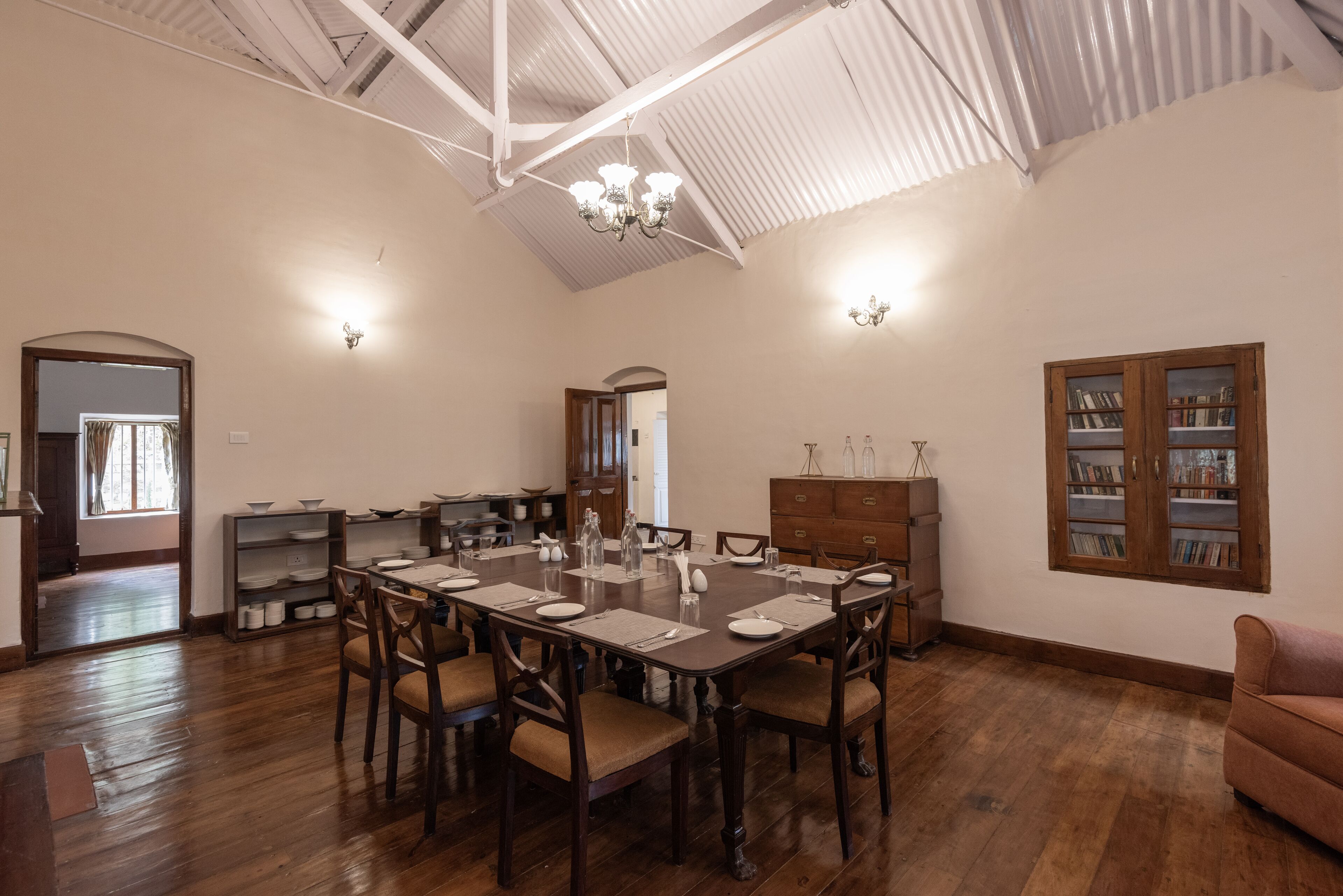 Un comedor con una gran mesa de madera dispuesta para seis personas, rodeada de sillas. Se aprecia una estantería con libros y objetos decorativos, un aparador de madera y una lámpara de araña en el techo.