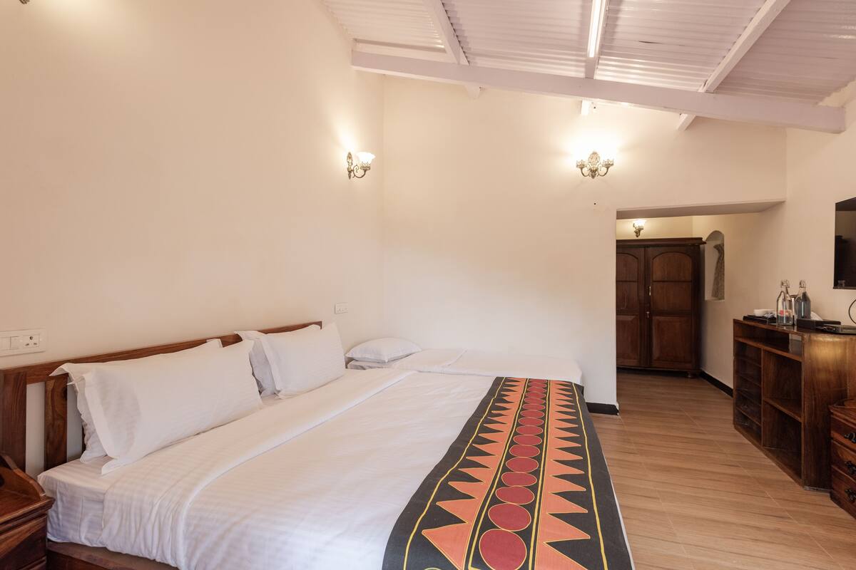 A bedroom with a large bed, wooden headboard, and a decorative blanket. There is a wooden wardrobe and a television on a wooden cabinet.