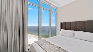 Une chambre avec une grande fenêtre donnant sur une plage, un lit avec du linge de lit blanc et une tête de lit grise.