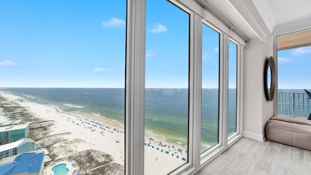 Une chambre d’hôtel située dans un immeuble de grande hauteur, dotée de grandes fenêtres du sol au plafond et offrant une vue sur une plage avec des parasols et une piscine.