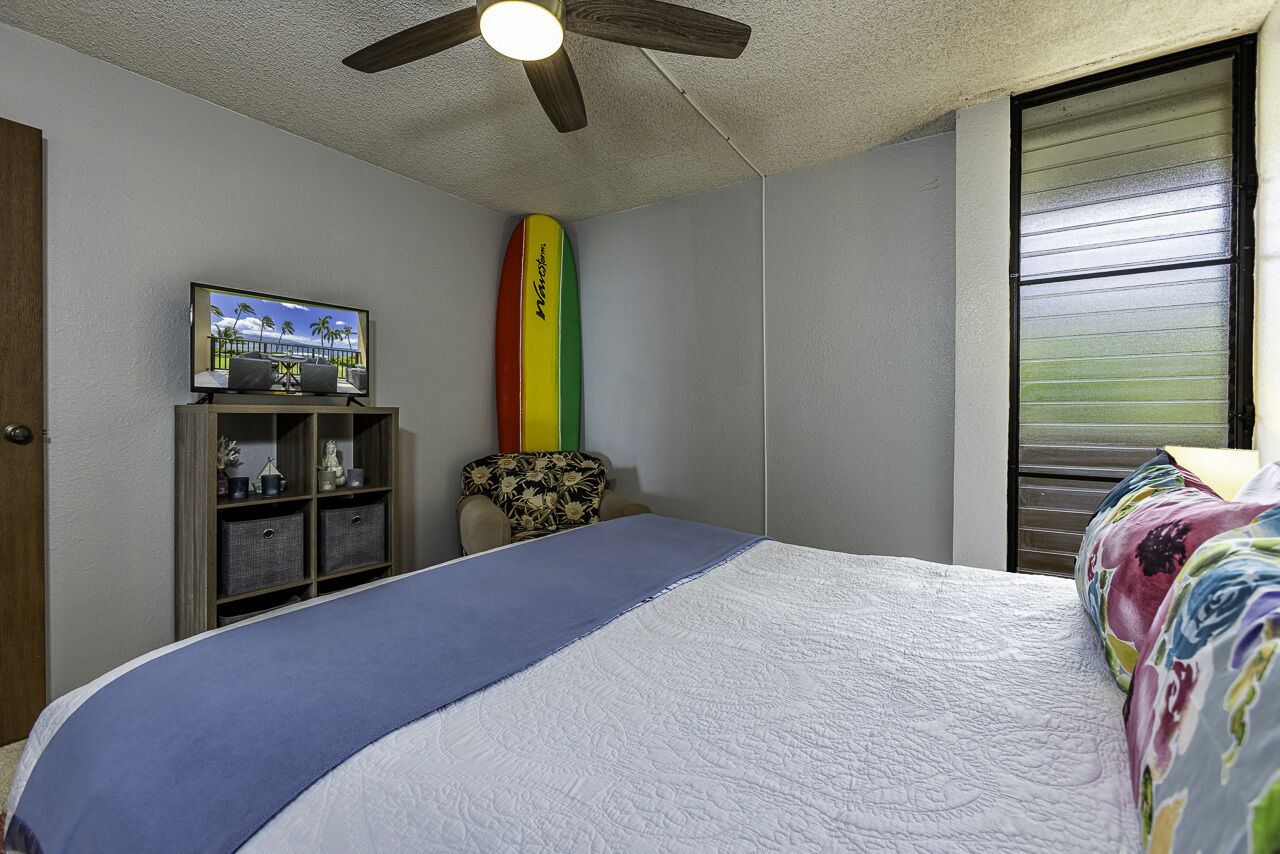 A bedroom with a bed, a ceiling fan, a surfboard, a TV, and a window with blinds.