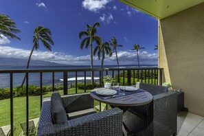 Un balcón con mobiliario de mimbre, una mesa dispuesta con platos y vasos, y vistas al océano y palmeras.