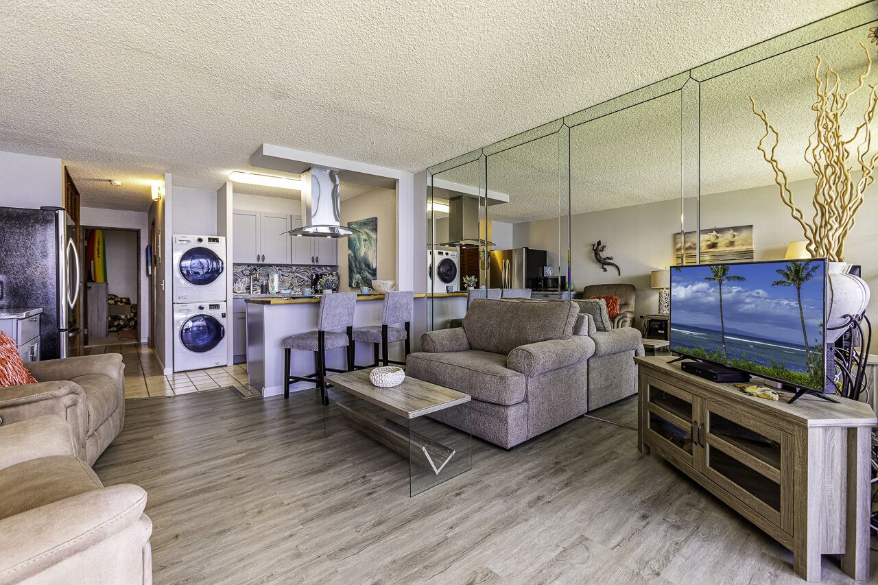 Una sala de estar moderna con un sofá, una mesa de centro y un televisor. Se ve una lavadora y una cocina al fondo.