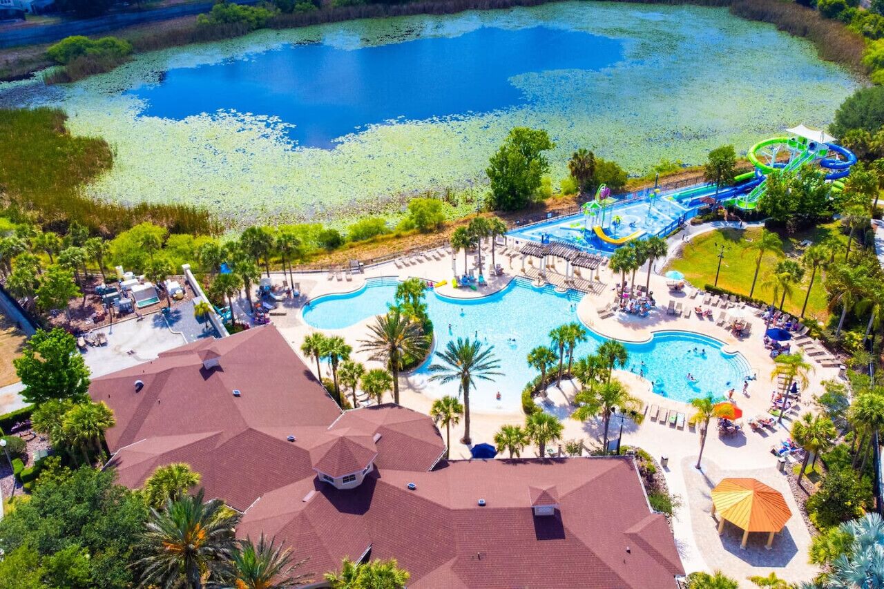 Vista aérea de un complejo turístico con múltiples piscinas, un parque acuático y un lago rodeado de palmeras.