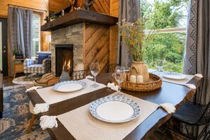 A dining area with a stone fireplace, a table set for two with plates and glasses, and a view of greenery outside.
