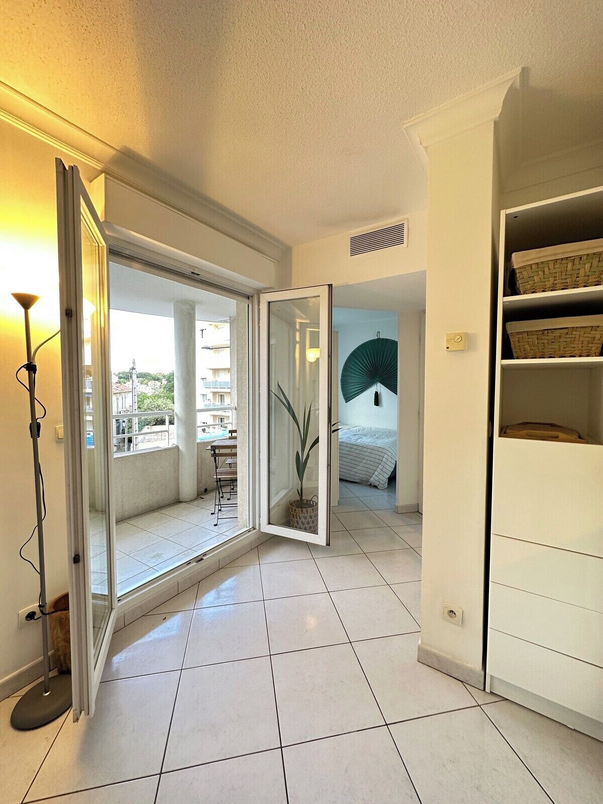 A room with a sliding glass door leading to a balcony, a bed with a green fan, and a white shelving unit with baskets.