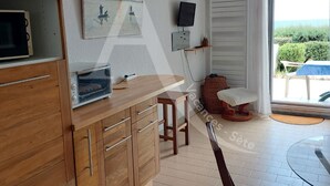Cozinha com armários de madeira, micro-ondas embutido, uma pequena mesa e uma cadeira. Há um quadro na parede e uma televisão sobre um suporte.