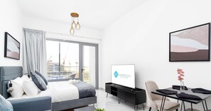 A modern hotel room with a bed, a desk, a chair, a TV, and a large window with a view of a construction site. - Elegant Studio in Oasis - 4 (Abu Dhabi)