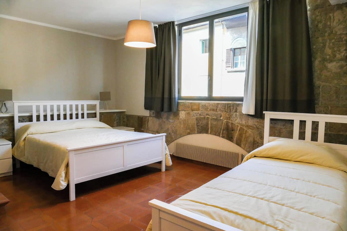 A room with two beds, a window with curtains, a stone wall, and a hanging light fixture.