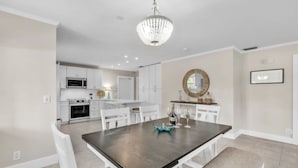 A modern kitchen with white cabinetry, a central island, and a dining area with a wooden table and white chairs. - Beached Inn 3 Bedroom Home by RedAwning (Holmes Beach)