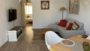 A modern living room with a grey sofa, a wooden dining table, and a flat-screen TV mounted on the wall.