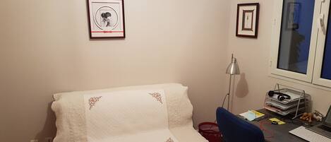 Ein kleiner, gemütlicher Raum mit einem Bett, einem Schreibtisch und einem Computer. An den Wänden hängen Bilderrahmen, es gibt eine Lampe und ein Fenster mit Blick.