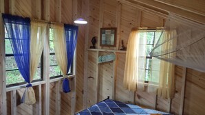 A wooden cabin interior with two windows, each with blue and yellow curtains. There is a bed with a blue quilt and a small shelf with a decorative item.