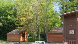 A wooden cabin with a 'Welcome' sign, a 'Parking' sign, and two hanging flower baskets.