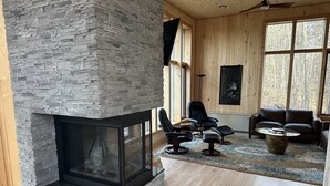 Un amplio salón con chimenea de piedra, suelo de madera y ventanas grandes que ofrecen vistas a árboles.
