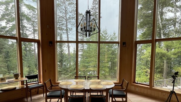 Una habitación amplia con una gran mesa de madera rodeada de sillas, ventanas amplias que ofrecen vistas al bosque, y una lámpara colgante.