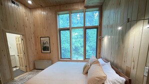 Un dormitorio con una cama grande, paredes de madera, una ventana con vistas a árboles y una puerta que da a otra habitación.
