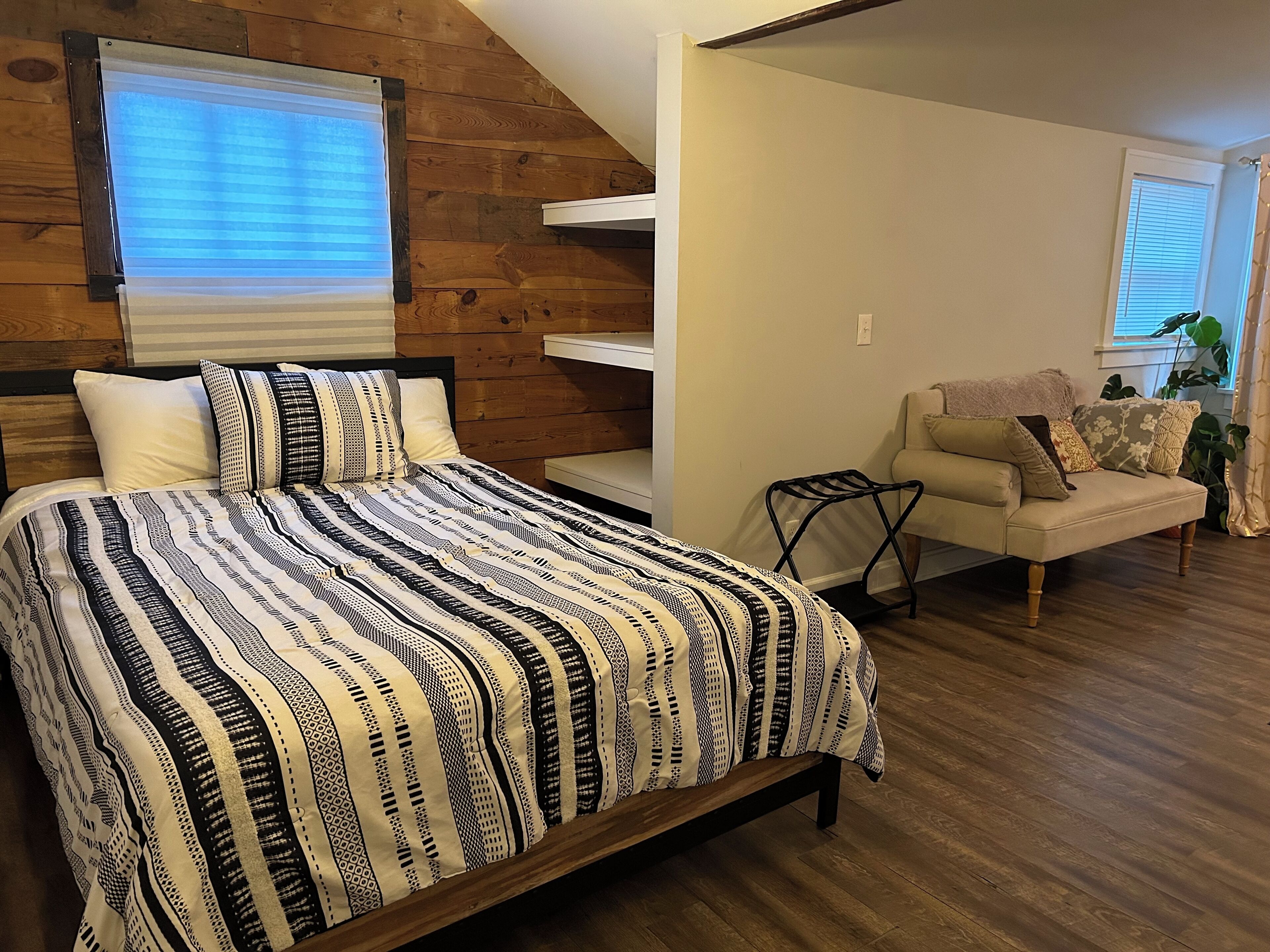 A bedroom with a bed, a sofa, a window with blinds, and a wooden ceiling.