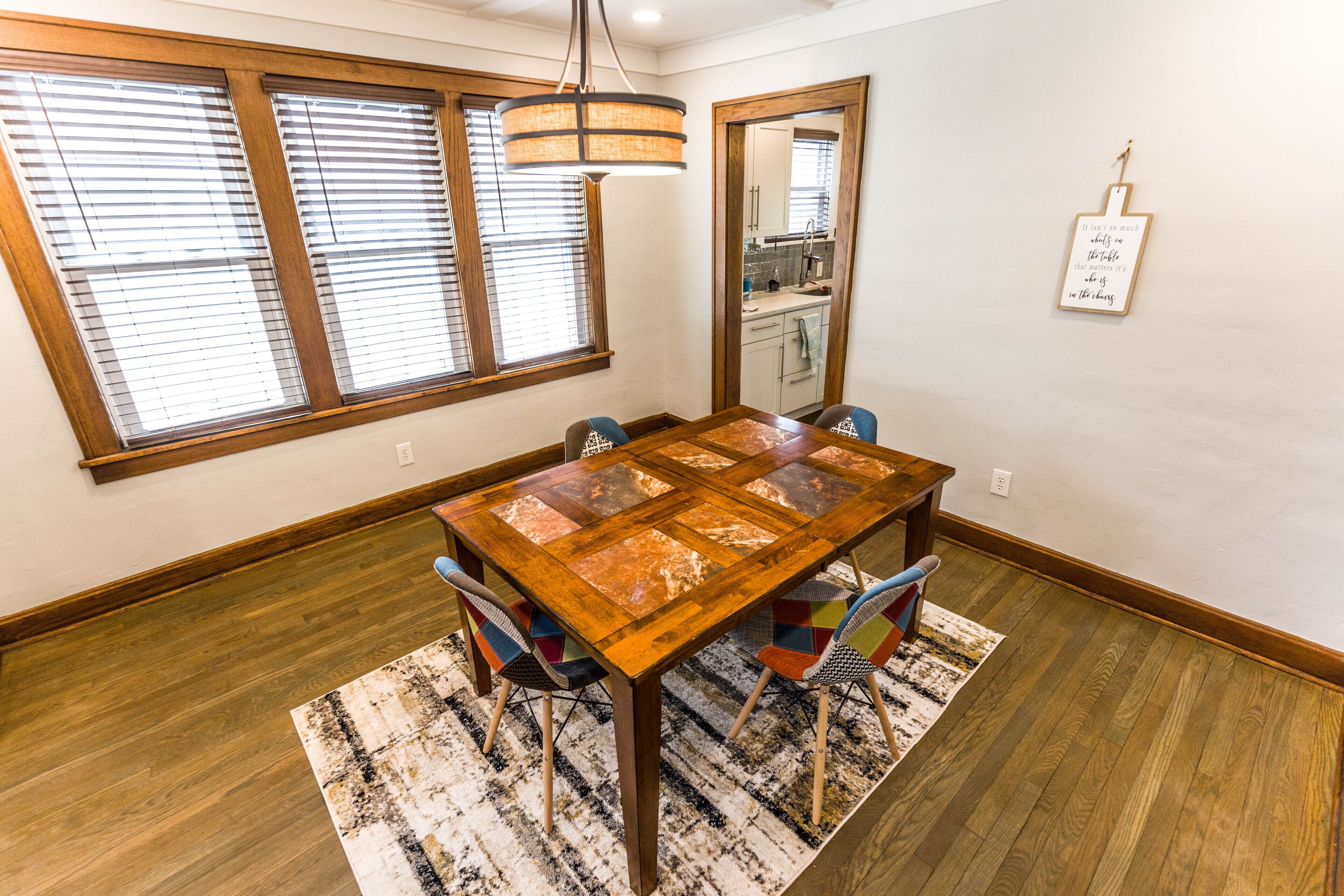 Un comedor con mesa de madera, sillas coloridas, alfombra estampada y ventanas amplias con persianas.
