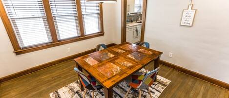 Un comedor con mesa de madera, sillas coloridas, alfombra estampada y ventanas amplias con persianas.