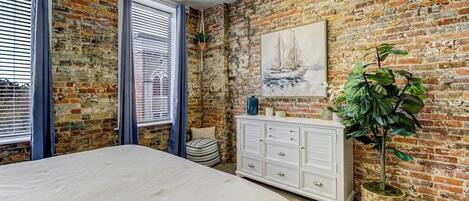 A bedroom with a brick wall, a bed, a dresser, and a window with curtains.