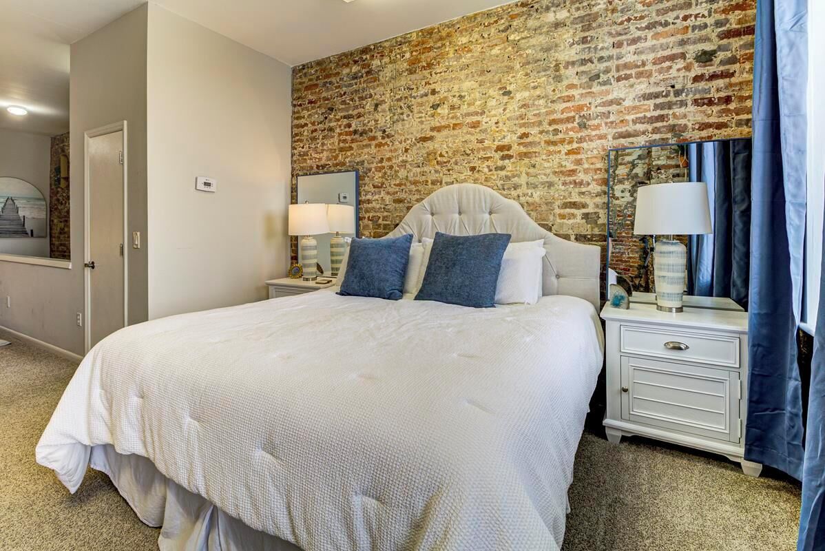 A hotel room with a large bed, white headboard, two nightstands with lamps, a brick wall, and a door to another room.