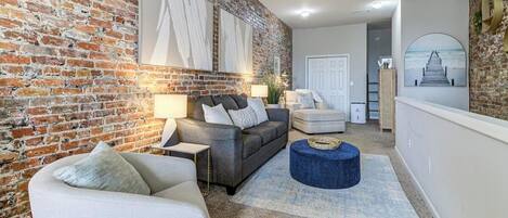 A living room with a brick wall, a grey sofa, a white armchair, and a blue ottoman.