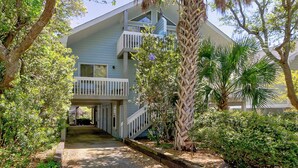 A two-story building with a balcony, surrounded by lush greenery and a palm tree. - Kure Beach Treehouse | Walk to Beach | Sleeps 8 (Kure Beach)