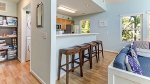 A kitchen with wooden cabinets, a refrigerator, and a window. There are three wooden stools and a bed with a patterned pillow. - Kure Beach Treehouse | Walk to Beach | Sleeps 8 (Kure Beach)