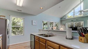 A kitchen with wooden cabinets, a stainless steel sink, and a window with blinds. - Kure Beach Treehouse | Walk to Beach | Sleeps 8 (Kure Beach)