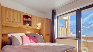 A bedroom with a wooden headboard, a large bed with multiple pillows, a nightstand, and a window with a mountain view. - Les Olympiades by Interhome (Val Thorens)