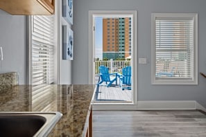 A kitchen with a granite countertop, a sink, and a door leading to a balcony with blue chairs and a view of a tall building.