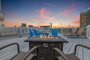 A rooftop deck with a fire pit, blue Adirondack chairs, and wicker lounge chairs.