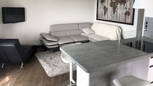 A modern living space with a grey sofa, a small dining table, a black chair, a white rug, and a mounted television. - Appartement Idéal Découverte Landes Pays Basques (Tarnos)