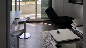 A modern living room with a sofa, a coffee table, and a TV mounted on the wall. There is a balcony with a view of greenery and a cityscape. - Appartement Idéal Découverte Landes Pays Basques (Tarnos)