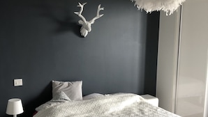 A bedroom with a dark wall, a white bedspread, a white lamp, and a white fluffy hanging light fixture. - Appartement Idéal Découverte Landes Pays Basques (Tarnos)