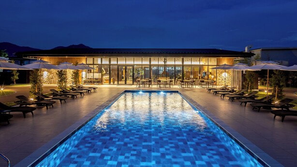 Une piscine avec des chaises longues et des parasols, un espace bar et un bâtiment doté de grandes fenêtres.