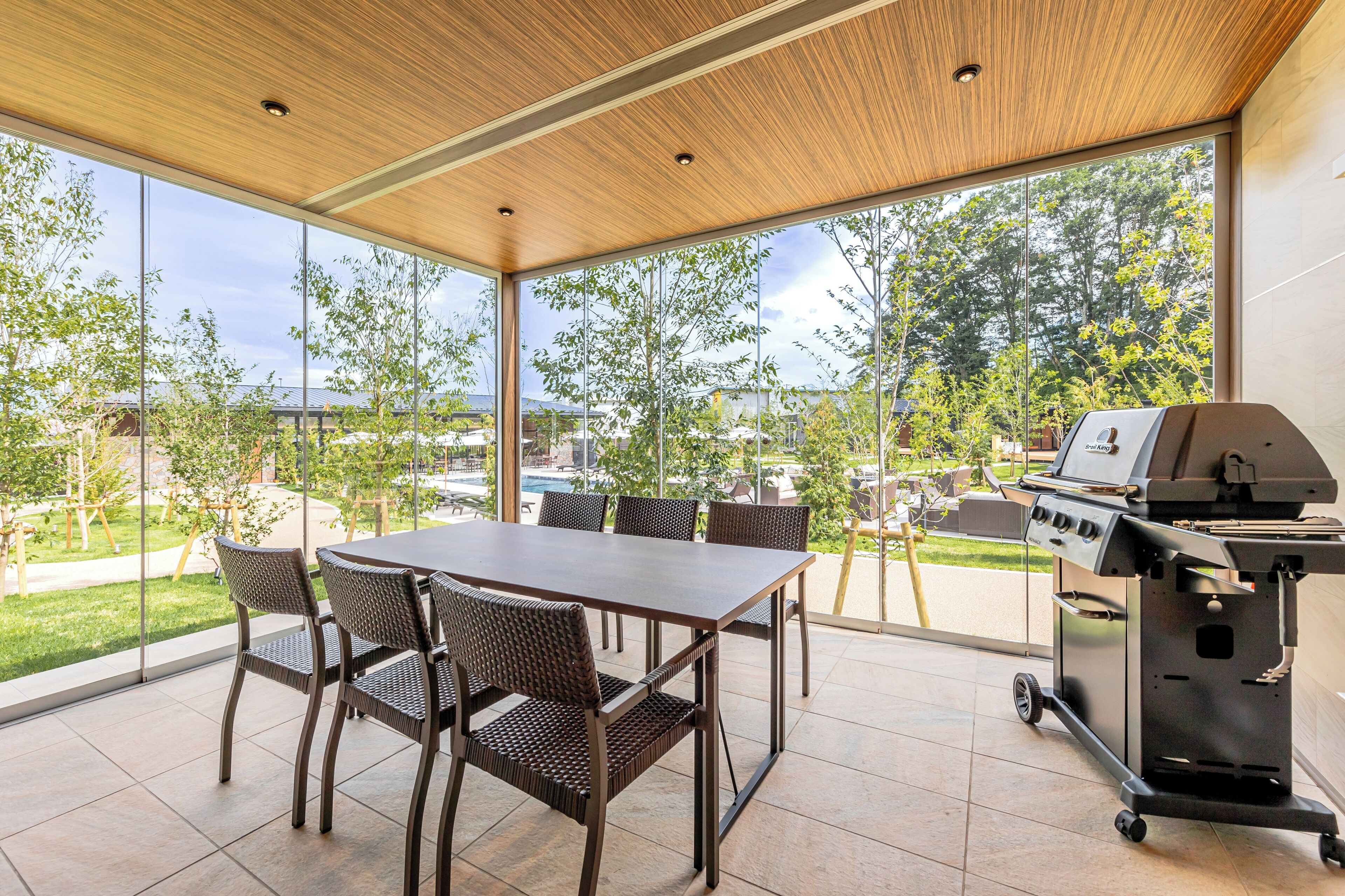 A patio with a table and chairs, a grill, and a view of a pool and greenery.