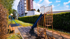 Ein Spielplatz mit Rutsche und Bank, umgeben von einer Hecke, mit einem Gebäude im Hintergrund.