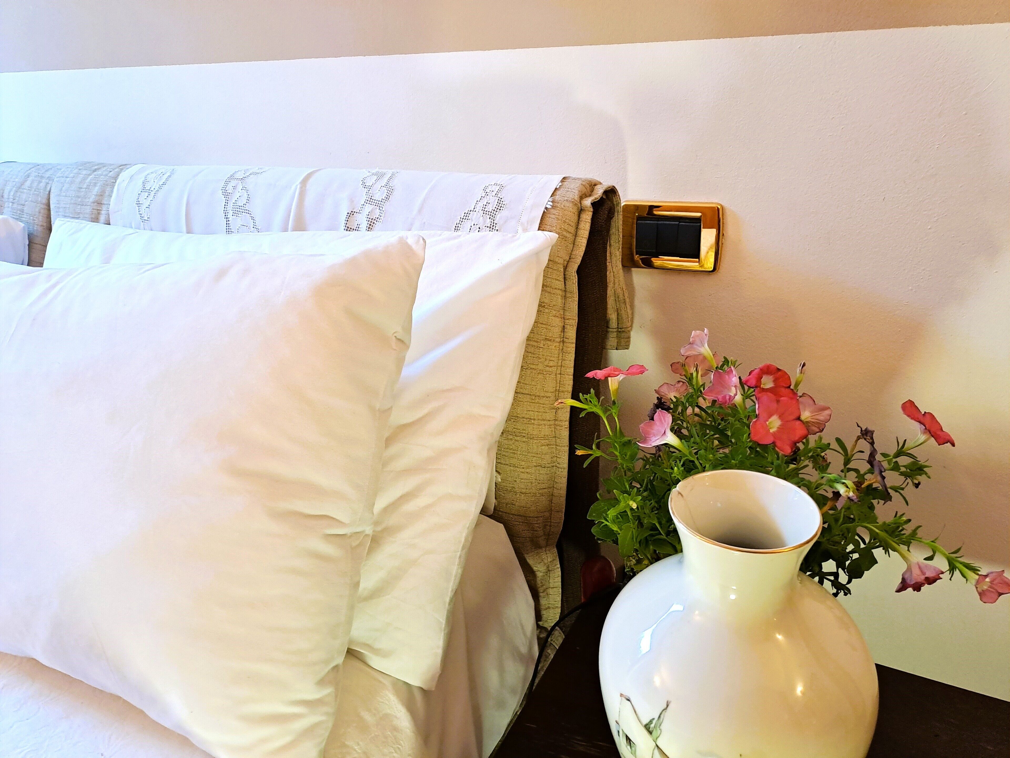 A neatly made bed with white linens, a small vase with flowers, and a nightstand with a clock.