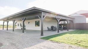 A covered patio area with outdoor seating and a grill, adjacent to a white building with windows. - COW PALACE 
Experience Rustic Charm and Tranquility at our Old Milk Barn.  (Preston)