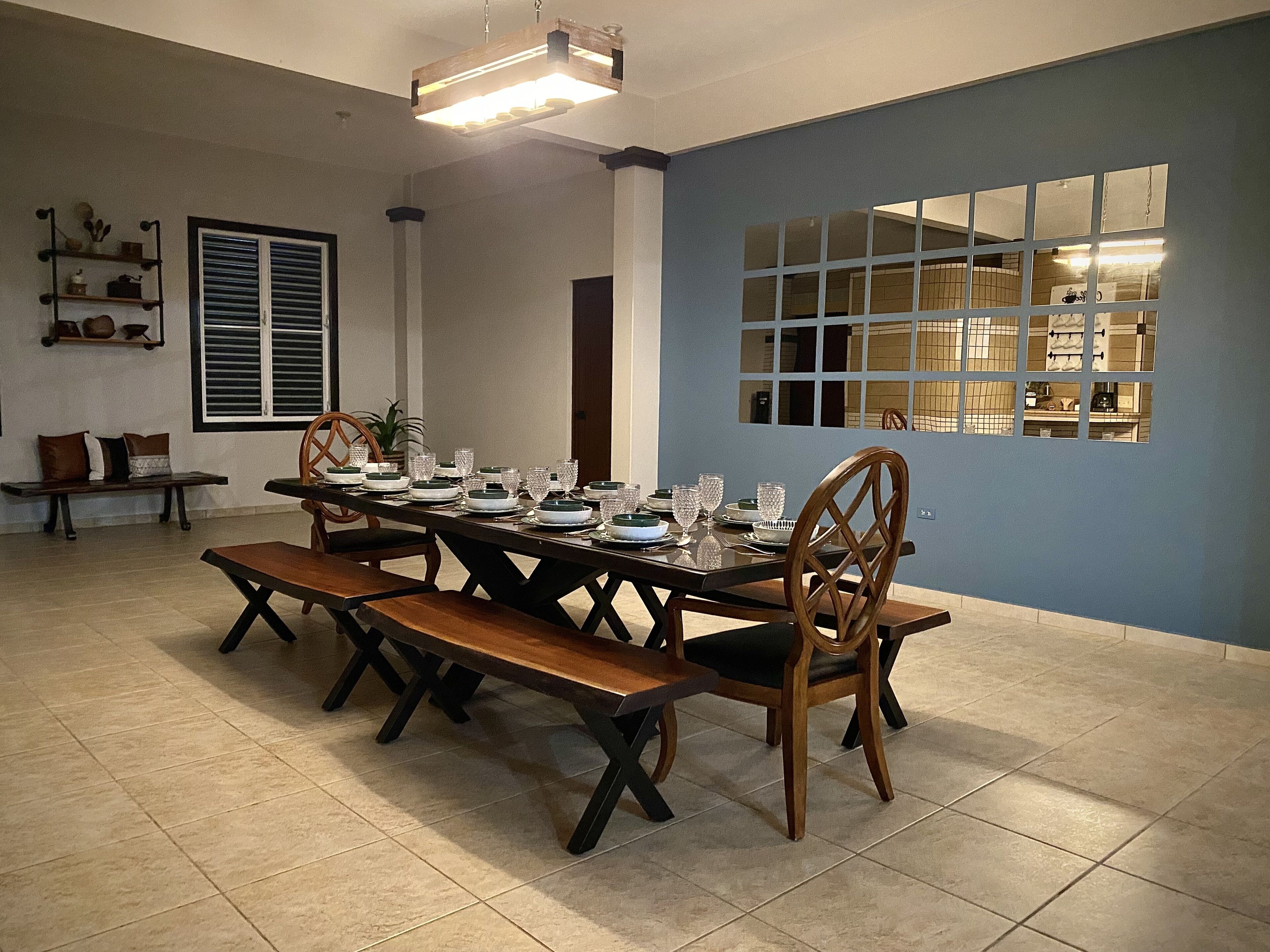 A dining area with a long table set for six, surrounded by wooden chairs. There is a bench to the left, a window with blinds, and a shelf with decorative items.