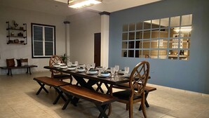 A dining area with a long table set for six, surrounded by wooden chairs. There is a bench to the left, a window with blinds, and a shelf with decorative items. - Private & spacious riverside home with HUGE pool, 10 mins to Rincon beaches! (Aguada)