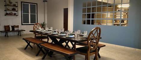 A dining area with a long table set for six, surrounded by wooden chairs. There is a bench to the left, a window with blinds, and a shelf with decorative items.