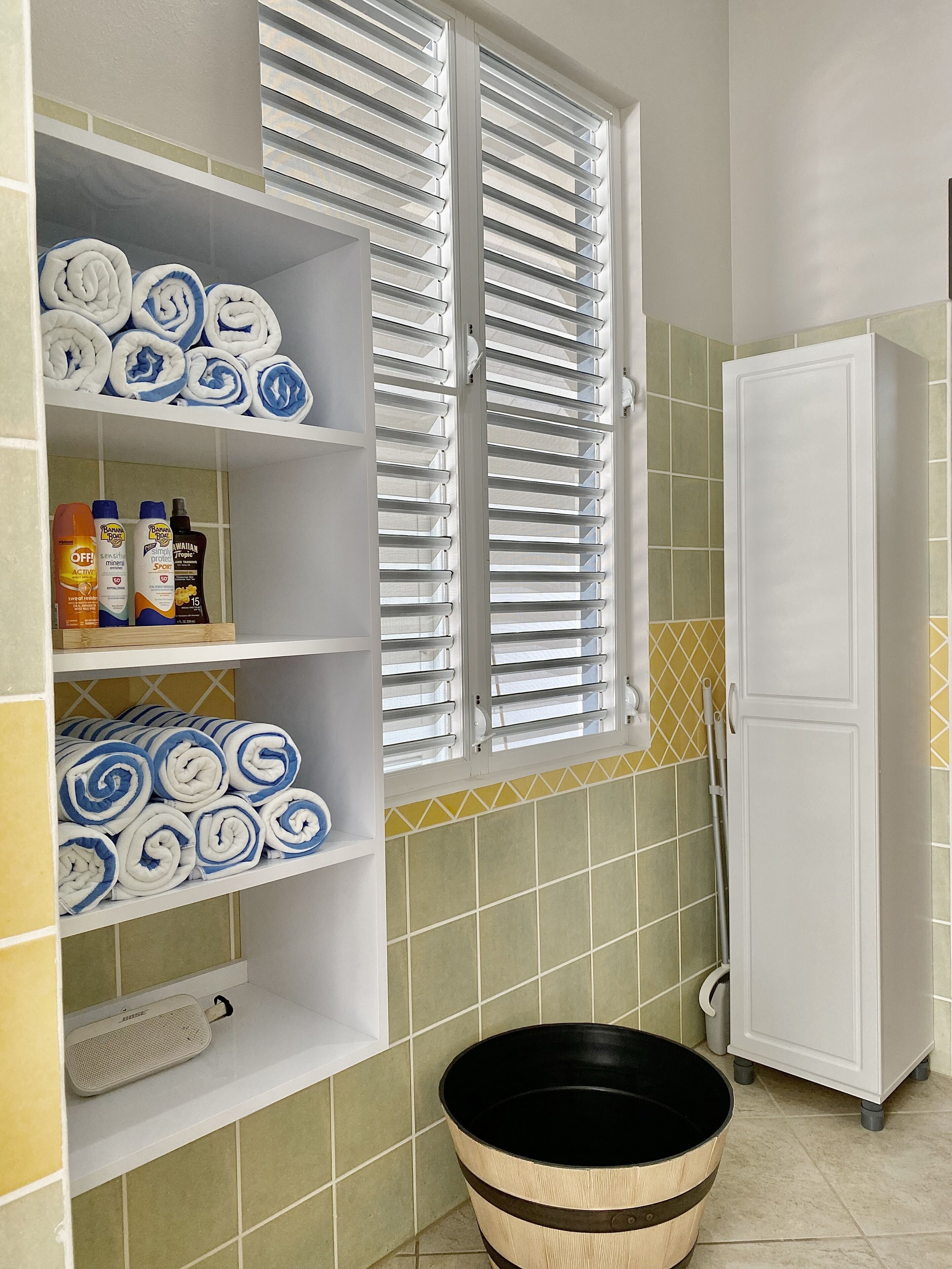 A bathroom with a shelf holding towels, toiletries, and a speaker. There is a window with blinds, a mop, and a cabinet.