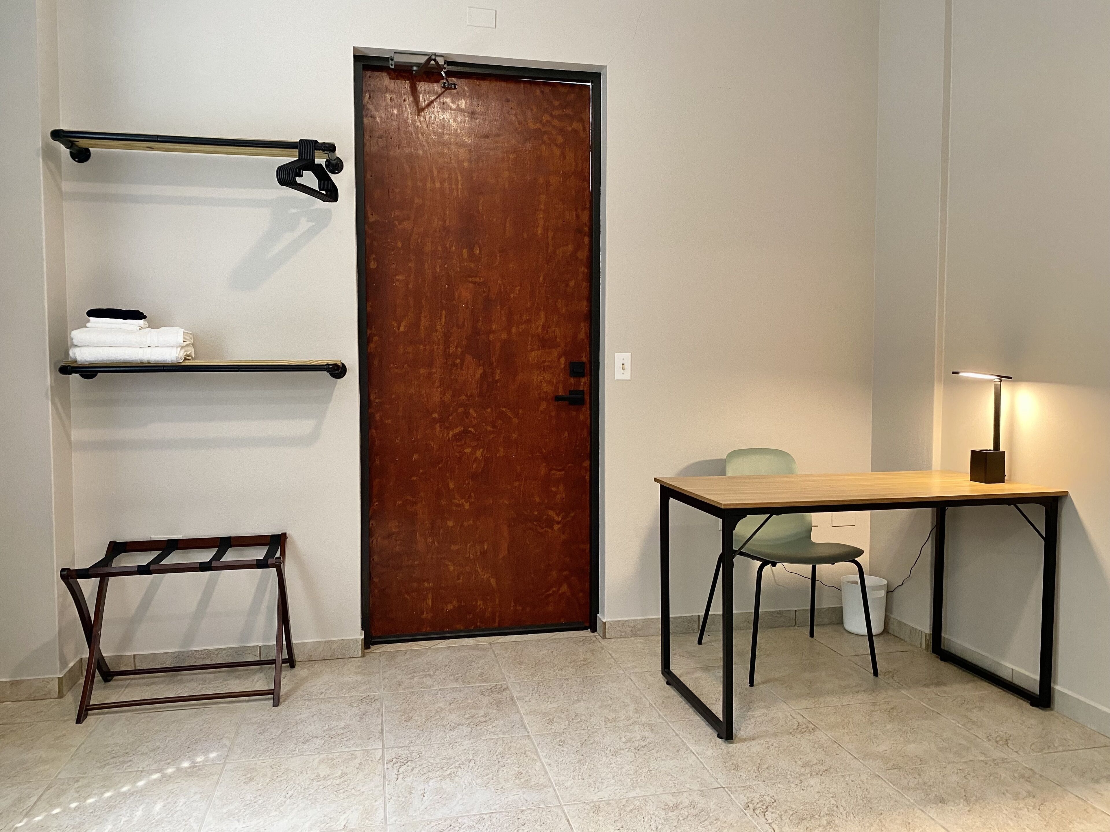 Una habitación con una puerta de madera, un perchero metálico con toallas, una mesita con una lámpara y una silla.