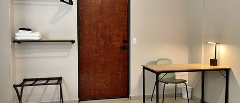 Una habitación con una puerta de madera, un perchero metálico con toallas, una mesita con una lámpara y una silla.