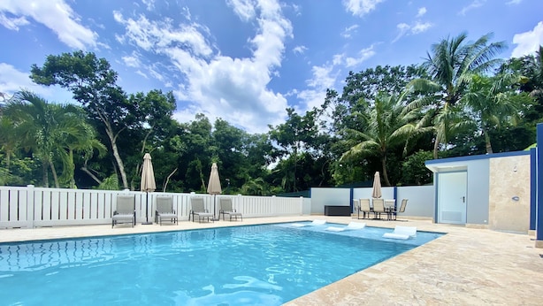A swimming pool with lounge chairs and umbrellas, surrounded by a white fence and palm trees. - Private & spacious riverside home with HUGE pool, 10 mins to Rincon beaches! (Aguada)